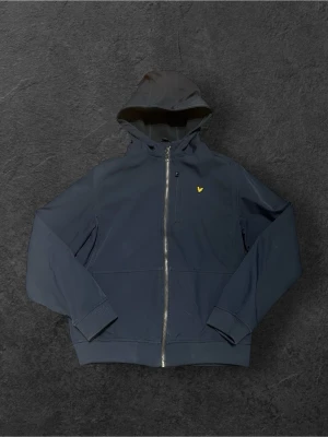 Mörkblå jacka från Lyle & Scott - Snygg mörkblå vindjacka från Lyle & Scott med huva och hel dragkedja framtill. Jackan har det klassiska gula fågelmärket på bröstet och är tillverkad i ett slitstarkt syntetmaterial. Perfekt för dig som vill ha en stilren och sportig look.