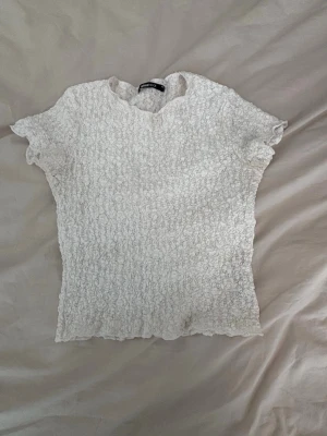 Vit blus - Supersöt vit blus med korta ärmar. Blusen har ett unikt, krusigt och texturerat tyg som ger en cool och lekfull känsla. Rund halsringning och stretchig passform som sitter snyggt på kroppen. Tröjan är kortare än vanliga T-shirts.