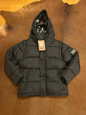 Svart pufferjacka från Burberry - Svart pufferjacka från Burberry med huva och klassisk Burberry-logga på ärmen. Jackan har quiltad design, dragkedja och knappar framtill samt fickor med dragkedja. Insidan av huvan har rutigt mönster i svart, vitt och rött. Perfekt för kalla dagar.