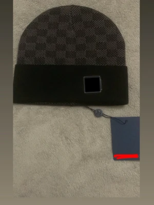 Svart och grå rutig mössa - Snygg mössa från okänt märke i svart och grått med klassiskt rutigt mönster. Mössan har en bred svart uppvikt kant och diskret logga på etiketten. Materialet är mjukt och värmande, perfekt för kyliga dagar