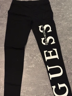 Svarta leggings från Guess - Svarta leggings från Guess med stor vit logotyp längs ena benet. Snygg och modern design med stretchigt material som sitter tight. Perfekt för dig som gillar att sticka ut med coola detaljer och stor branding. Storlek Xs