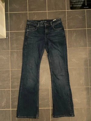 mörkblå bootcut jeans - Snygga mörkblå jeans med klassisk bootcut passform. Jeansen har fem fickor, bälteshällor och en brun läderpatch bak i midjan. Tillverkade i slitstarkt denim med raka ben och subtila kontrastsömmar. Perfekta för dig som gillar retrovibbar och tidlös stil.