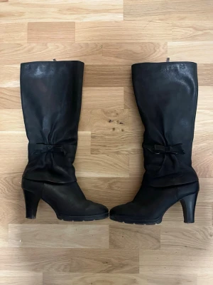 Svarta stövlar med rosett och klack - Snygga svarta stövlar i skinn med hög klack och rund tå. Stövlarna har en dekorativ rosett på sidan