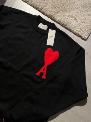 Svart stickad tröja från Ami Paris (trade bara) - Svart stickad tröja från Ami Paris med rund hals och långa ärmar. På bröstet finns ett stort rött hjärta med bokstaven A, som är typiskt för Ami. Tröjan har ribbade muddar vid ärmslut och nederkant, vilket ger en snygg och clean look.