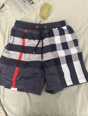 Burberry shorts - Snygga Burberry shorts med klassiskt rutigt mönster i mörkblått, vitt, grått och en röd detalj. De har elastisk midja med snörning och är perfekta för sommaren. Materialet är lätt och känns skönt mot huden. Ikonisk design som sticker ut.