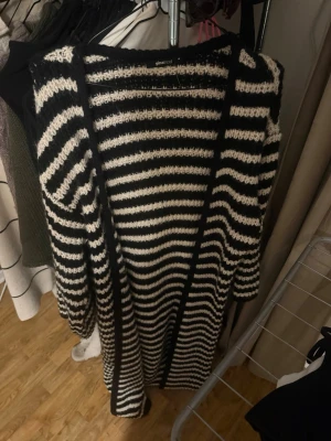 Svartvit randig stickad kofta Gina Tricot - Säljer en lång, stickad kofta från Gina Tricot i storlek M. Koftan har ett snyggt svartvitt randigt mönster och är öppen framtill utan knappar. Perfekt att slänga över en t-shirt för en chill och trendig look. Långa ärmar och mjukt material.