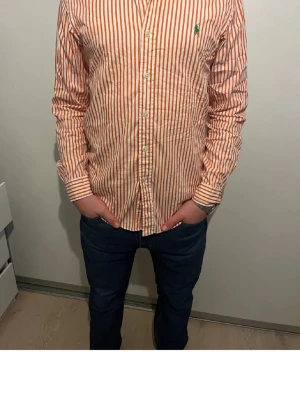 Randig skjorta Polo Ralph Lauren M - Orange och vit randig skjorta från Polo Ralph Lauren i slim fit. Klassisk button-down krage, långa ärmar och grönt broderat logomärke på bröstet. Skjortan är i bomull och har knäppning framtill.
