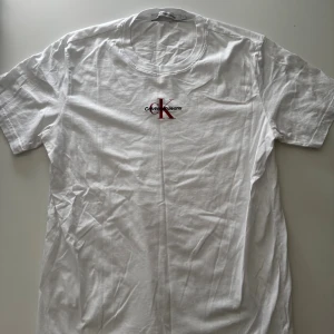 Vit Calvin Klein Jeans t-shirt L - Vit t-shirt från Calvin Klein Jeans i storlek L. Klassisk modell med rund hals och korta ärmar. Framtill finns en snygg CK-logga i rött och svart. Tillverkad i mjuk bomull som är skön mot huden. Perfekt för en clean och stilren look.