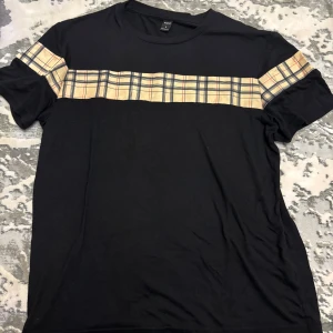 Svart t-shirt med rutig detalj - Svart t-shirt från Brave Soul med korta ärmar och en beige-rutig panel över bröstet och axlarna. Klassisk passform och rund halsringning. Perfekt för dig som gillar streetstyle med en twist.