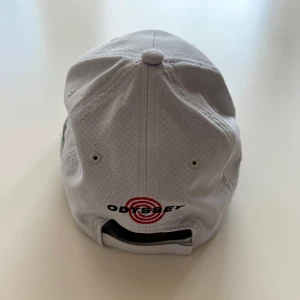 Vit Callaway keps med broderi - Snygg vit keps från Callaway med broderade loggor i svart, grönt och gult. Kepsen har texten 'Chrome Soft', 'Apex', 'Epic Flash' och 'Odyssey' broderat runt om. Tillverkad i ventilerande syntetmaterial med justerbar rem bak. Perfekt för golf eller sportiga dagar.