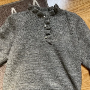 Grå stickad tröja Holly & Whyte XS - Mysig grå stickad tröja från Holly & Whyte med glittriga inslag, volangkrage och pärlknappar framtill. Tröjan har lång ärm och ribbade muddar. Perfekt för dig som gillar detaljer och en klassisk look.