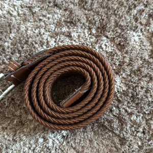 Brunt flätat bälte med silverfärgat spänne bvaldi  - Snyggt brunt bälte med flätad design och silverfärgat spänne. Bältet har detaljer i brunt skinn vid spännet och ändarna, vilket ger en stilren och klassisk look. Perfekt för att lyfta vilken outfit som helst med en touch av vintage-vibe.