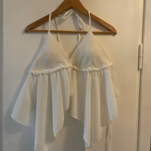 Vit topp med knyten och helt öppen rygg - Säljer en vit topp som man knyter i nacken och i ryggen. Toppen har en flowy, asymmetrisk nederdel med helt öppen rygg och är tillverkad i ett lätt tyg som ger en luftig känsla. Köpt i Spanien! Passar alla i storleken eftersom man knyter den utefter hur man vill ha den!