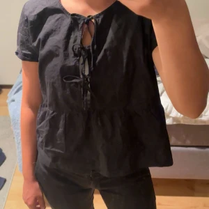 Svart blus med knyt fram - Supergullig blus från Gina med korta ärmar och knytband framtill. Modellen har en lätt volang nertill och rundad halsringning. Toppen är i storlek s🥰 