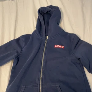 Mörkblå hoodie från Levi's med dragkedja - Säljer en mörkblå hoodie från Levi's med klassisk röd logga på bröstet. Tröjan har huva, dragkedja framtill och två fickor. Perfekt för dig som gillar en enkel och stilren look. Materialet är mjukt och skönt, troligen bomullsmix.