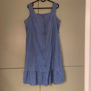 Blå prickig klänning - Supersöt blå klänning med vita prickar från Danielle Philipe. Klänningen har breda axelband, knappar framtill och en volang längst ner. Tillverkad i 100% bomull, perfekt för varma dagar💘