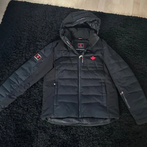 Svart pufferjacka från Canada Snow - Säljer en svart pufferjacka från Canada Snow med röd lönnlöv-detalj på bröstet och logga på ärmen. Jackan har huva, dragkedja framtill och quiltad design. Perfekt för kalla dagar och har en clean, sportig look. perfekt kalla vinterdagar