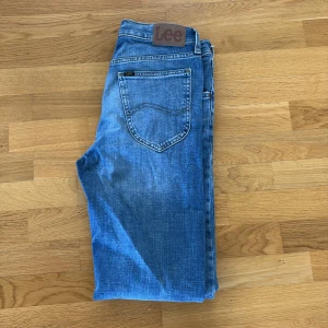 Blå Lee jeans straight fit - Klassiska blå jeans från Lee med raka ben och normal passform. Jeansen har fem fickor, bälteshällor och Lee-logga på bakfickan. Tillverkade i slitstarkt bomullsjeansmaterial med snygga slitningar och ljusare partier framtill.29/32.