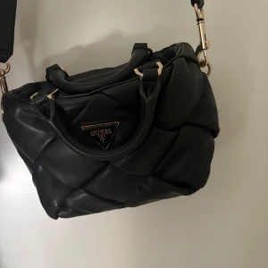 Svart quiltad axelväska från Guess - Snygg svart axelväska från Guess med quiltad design och guldfärgade detaljer. Väskan har både handtag och justerbar axelrem, samt den klassiska Guess-loggan framtill. Tillverkad i skinnimitation som ger en lyxig känsla. Säljer på grund av att jag har växt ifrån den. Pris kan diskuteras vid snabb köp