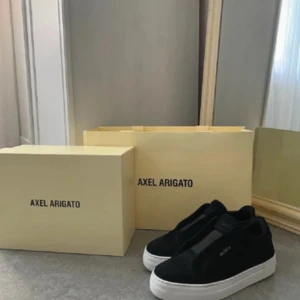 Svarta sneakers från Axel Arigato - Snygga svarta sneakers från Axel Arigato med minimalistisk design och vit platt sula. Skorna har rund tå och är tillverkade i mocka med diskret logga på sidan. Perfekta för en clean och stilren look. Använt några gånger bara ser ut som nya 