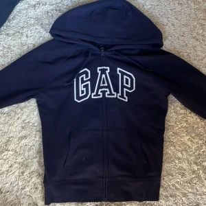 Mörkblå hoodie från GAP med dragkedja - Mörkblå hoodie från GAP med stor vit logga framtill. Tröjan har huva med snörning, dragkedja och två fickor. Tillverkad i en mjuk blandning av bomull och polyester. Perfekt för en chill och avslappnad stil. Det är bara att fråga om du vill ha fler bilder😊 Andvänd helst inte köp direkt utan skriv först tack 😊 det är en dammodell