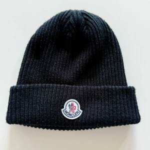 Svart ribbad mössa från Moncler - Svart ribbstickad mössa från Moncler med klassisk uppvikt kant och broderad logotyp framtill. Perfekt accessoar för kalla dagar och en stilren detalj till din outfit.