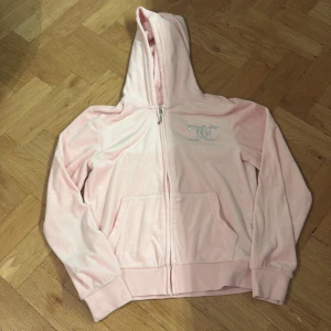 Rosa Juicy Couture hoodie i velour - Superfin ljusrosa hoodie från Juicy Couture i mjuk velour med glittrande strasstenar på bröstet och stor Juicy Couture-text på ryggen. Tröjan har huva, dragkedja och två fickor framtill. Perfekt för dig som älskar Y2K-stil!