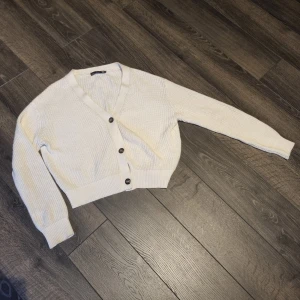 Beige croppad kofta med knappar - Superfin beige croppad kofta med v-ringning och stora knappar framtill. Stickad i mjukt material och har långa ärmar. Perfekt att slänga över en topp för en chill look.