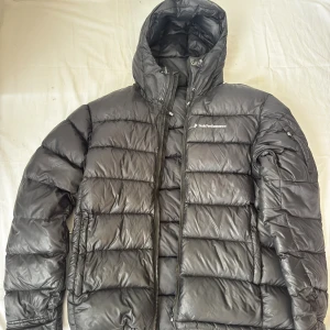 Svart dunjacka Peak Performance storlek L - Säljer en svart dunjacka från Peak Performance i storlek L. Jackan har huva, tvåvägsdragkedja och flera fickor. Klassisk pufferstil med breda kanaler och logga på bröstet. Perfekt för kalla dagar och enkel att matcha med allt.