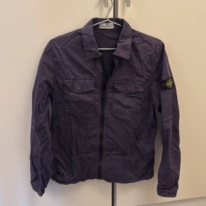 Overshirt från Stone Island  - Säljer en snygg overshirt från Stone Island med klassisk logga på ärmen. Jackan har två bröstfickor med lock, dragkedja framtill och en stilren krage. Perfekt för dig som vill ha en clean och trendig look. Jackan är ungefär xs-s. Priset är inte hugget i sten!