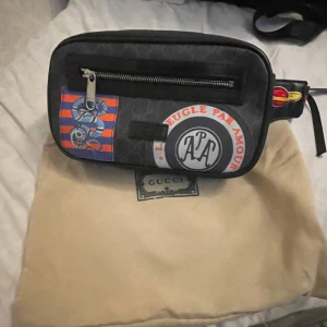 Gucci svart axelväska med patchar - Säljer en svart axelväska från Gucci med coola patchar och tryck, bland annat ormmotiv och texten 'L'aveugle par amour'. Väskan har en dragkedja framtill och justerbar rem i rött och svart. Materialet är skinn och detaljerna är i silverfärgad metall.