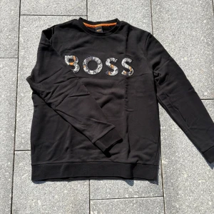Svart sweatshirt från Hugo boss  med tryck - Svart sweatshirt från Hugo boss  med stort, mönstrat BOSS-tryck i vitt, grått och orange framtill. Tröjan har rund halsringning med orange detalj på insidan och långa ärmar. Perfekt för en cool och avslappnad stil.