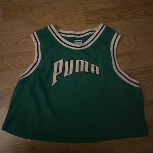 Grön croppad linnetopp från Puma - Sportig grön linnetopp från Puma med vita och svarta ränder längs halsringning och ärmhål. Croppad modell med stort Puma-tryck framtill. Perfekt för en avslappnad och cool streetstyle.