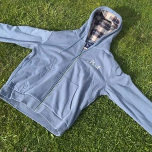 Blå hoodie från Leon Marcel  - Säljer en blå hoodie från Leon Marcel med dragkedja och huva. Insidan av huvan har ett rutigt foder i beige, rött och blått. Tröjan har fickor framtill och snörning i huvan. Perfekt för kyliga dagar och chill vibes. Den är xl men sitter som en large