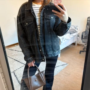 Svart oversized jeansjacka - Säljer en svart oversized jeansjacka med klassisk krage och silverfärgade knappar. Jackan har två bröstfickor med lock och knapp samt raka sömmar. Perfekt för dig som gillar en chill och avslappnad stil.