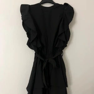 Volangklänning med shorts och knytband - Svart klänning med volangdetaljer på axlarna och knytband i midjan. Klänningen har ärmlös design och en liten öppning med knapp i nacken. Det är ett par inbyggda shorts.