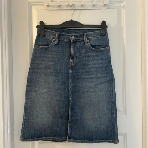 Levi's jeanskjol med slits, stl 26 - Snygg jeanskjol från Levi's i klassisk blå denim. Kjolen har fem fickor, hällor för bälte och en liten slits framtill för extra rörelsefrihet. Rak modell som slutar strax ovanför knät. Perfekt till sneakers eller boots.