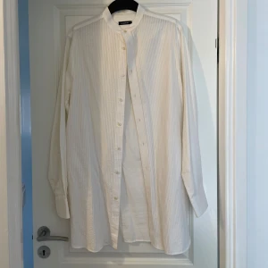 J.Lindeberg Vit henley-skjorta från  - Vit henley-skjorta från J.Lindeberg med ståkrage och diskreta vertikala ränder. Skjortan har knappar framtill och är långärmad. Tillverkad i bomull för en skön och luftig känsla. Perfekt för dig som gillar stilrena och moderna plagg.