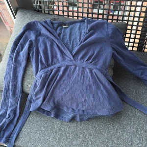 Blå omlottopp från Gina Tricot XS - Supersnygg blå omlottopp från Gina Tricot i storlek XS. Toppen har långa ärmar, v-ringning och knytband i midjan. Materialet är lätt och har en lite skrynklig struktur som ger en avslappnad vibe.