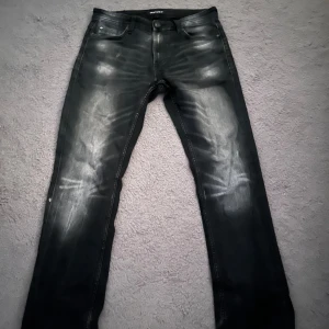 Replay svarta jeans med slitningar - Säljer ett par svarta Replay jeans med snygga slitningar och tvättade detaljer på benen. Jeansen har klassisk femficksdesign, raka ben och normal passform. Perfekta för dig som gillar en lite ruffare streetstil.