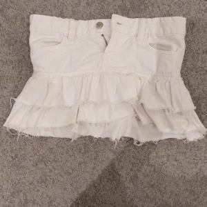 Vit volangkjol från Gina Tricot - Supersöt vit kortkjol från Gina Tricot med flera lager volanger och råa kanter för en trendig look. Kjolen har jeansfickor fram och bälteshällor, perfekt att styla med en cool topp eller hoodie.