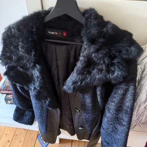 Svart pälsjacka från aviatRyk - Snygg svart pälsjacka med bred krage och bälte i midjan från aviatRyk. Jackan har glansig finish, stora knappar framtill och detaljer i skinnimitation. Perfekt för dig som vill sticka ut med en lyxig vibe.