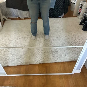 Ljusblå bootcut jeans med slitning - Säljer ett par ljusblå jeans med bootcut passform och en snygg sliten detalj vid ena knät. Jeansen har klassisk femficksdesign och är tillverkade i mjukt denimtyg. Dom är uppsydda längst ner för de var för långa. Så är du runt 166cm så passar dom perkekr för dig!! 