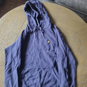 Marinblå hoodie från Lyle & Scott - Snygg marinblå hoodie från Lyle & Scott med klassisk gul logga på bröstet. Tröjan har huva med snörning och en stor magficka framtill. Perfekt för dig som gillar en avslappnad och stilren look.