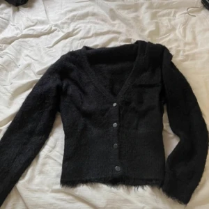 Svart fluffig kofta från Vero Moda - Mysig svart kofta från Vero Moda i storlek S. Koftan är stickad med fluffig känsla, har v-ringning och knäpps med svarta knappar framtill. Perfekt för lager-på-lager och passar till många olika outfits.