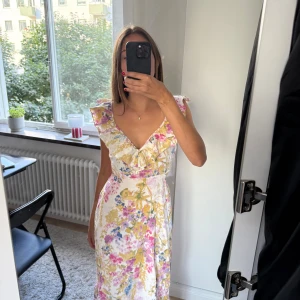 Blommig långklänning med volang - Superfin långklänning med blommigt mönster i rosa, gult och blått på vit botten. Djup v-ringning med volangdetaljer och breda axelband och öppen rygg. Använt 1 gång på midsommar. 