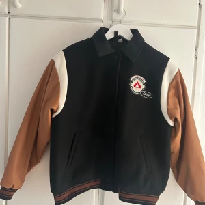 Svart och brun bomberjacka Outdoor Camper - Snygg svart bomberjacka med bruna ärmar och vita detaljer från Outdoor Camper. Jackan har broderade patches framtill och stort tryck med '1990' på ryggen. Klassisk college-stil med ribbade muddar och knappar framtill. Perfekt för dig som gillar retrovibbar.