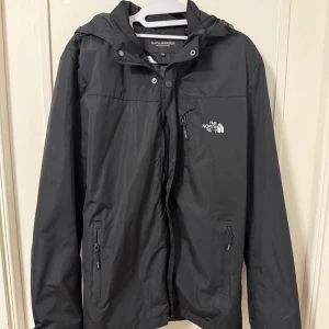 Svart vindjacka från The North Face - Svart vindjacka från The North Face med huva och dragkedja framtill. Jackan har två sidofickor med dragkedja och en bröstficka. Perfekt för blåsiga dagar och enkel att matcha med allt. Klassisk logga på bröstet.