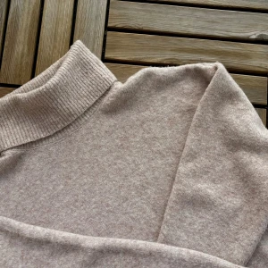 Beige/Sandfärgad stickad tröja - Mjuk polotröja i en beige/sand färg med ribbad och hög krage. Tröjan är stickad i en mjuk ullmix och har lång ärm, perfekt för hösten och våren. 