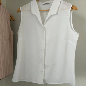 Vit ärmlös skjorta från BHS - Vit ärmlös skjorta från BHS med klassisk krage och knappar framtill. Skjortan har en lätt strukturerad yta och rak passform, perfekt för en clean och fräsch look. Materialet känns svalt och luftigt, vilket gör den nice till varmare dagar.